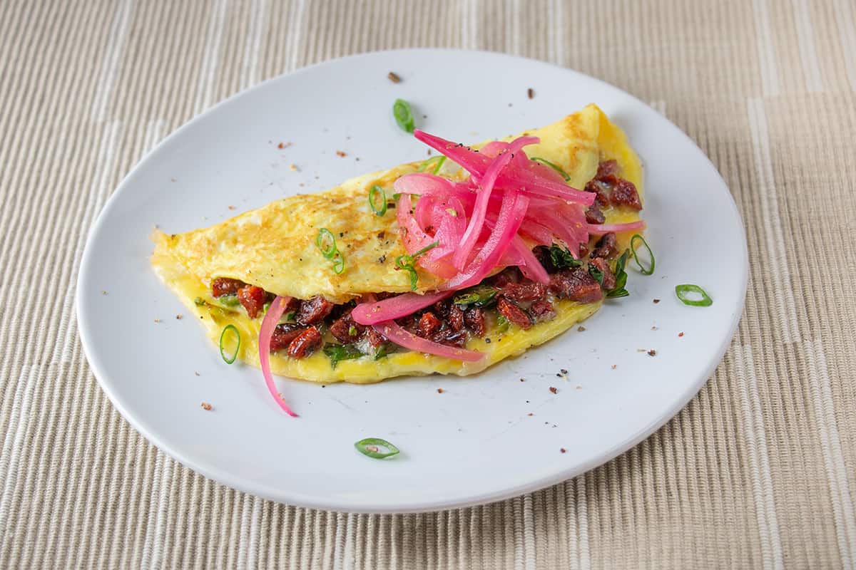 Omelette filled with a red-coloured sausage and topped with pink pickled onions.