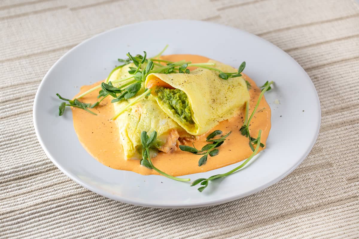 White plate covered with an orange sauce with an omelette stuffed with salmon and peas.
