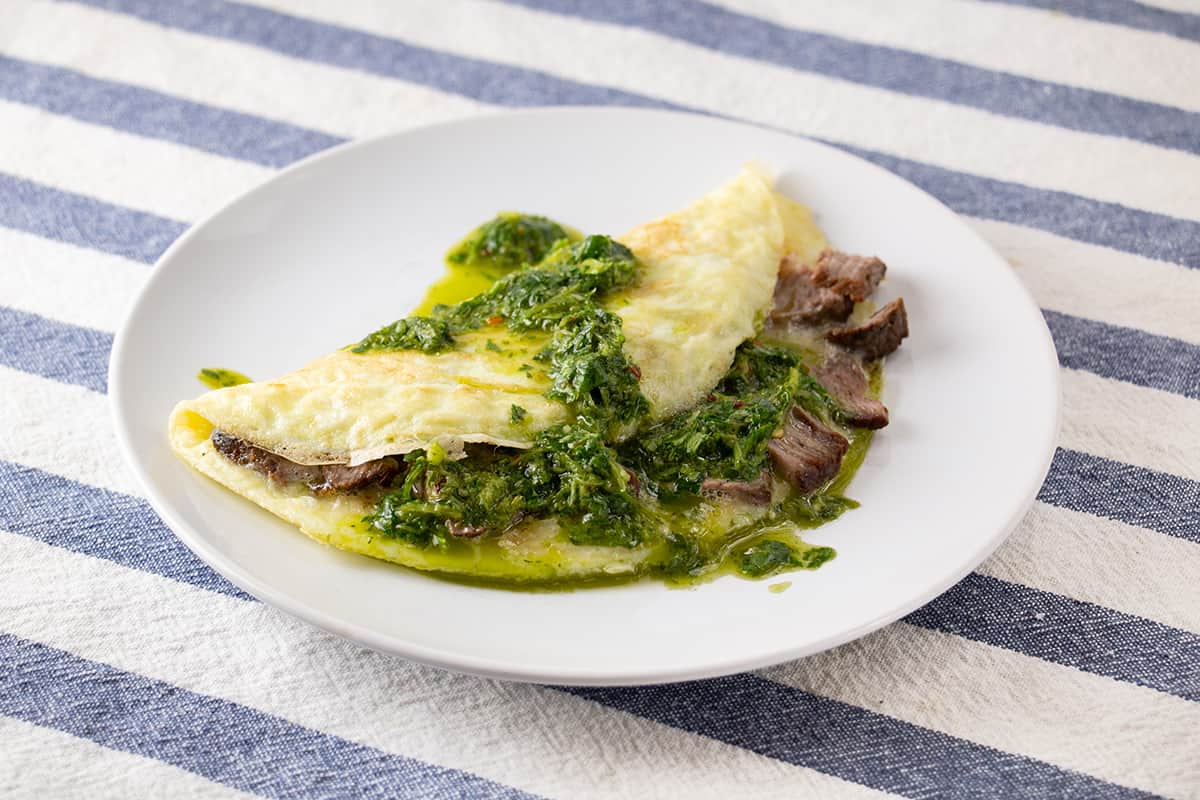 White plate with an omelette filled with steak and topped with a green sauce.