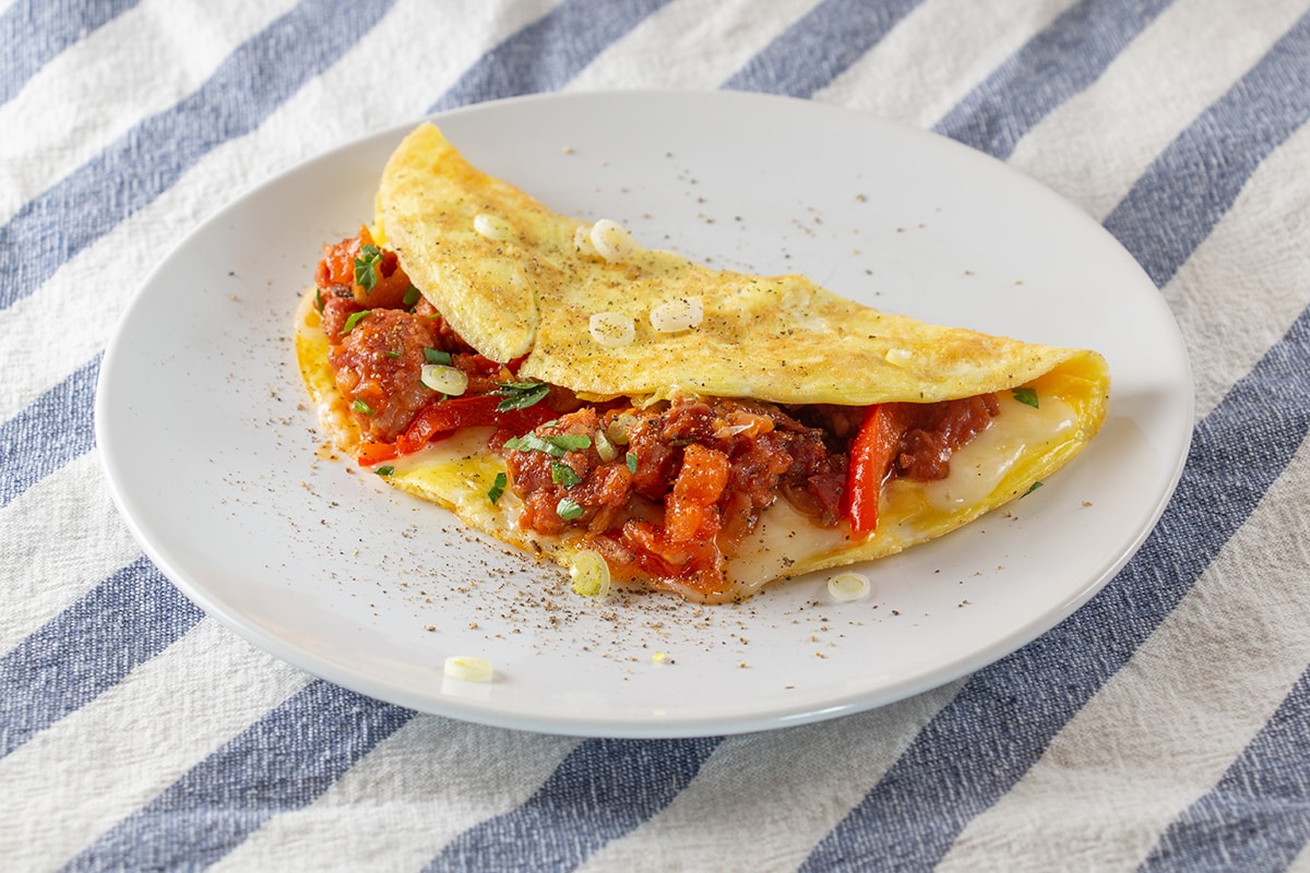 Omelette on a white plate, filled with sausage, peppers and a red sauce.