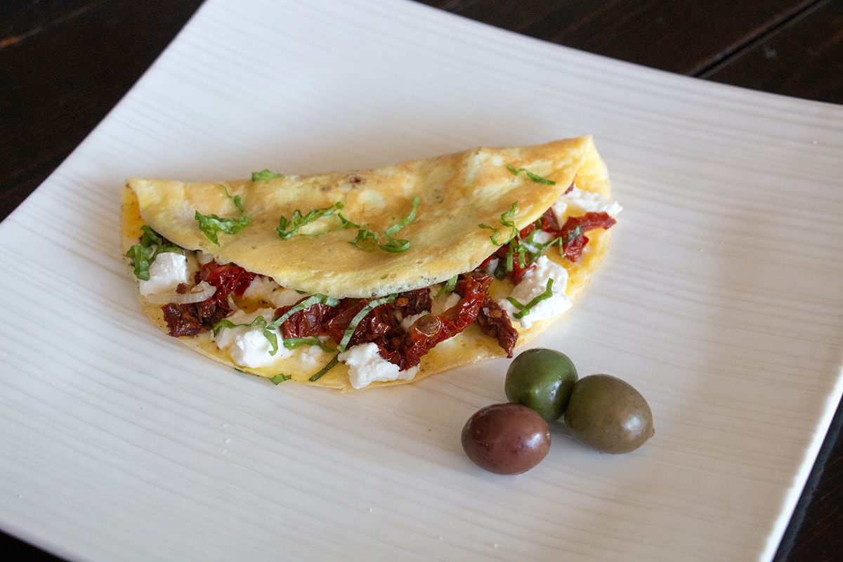 White plate with an omelette filled with goat cheese, tomatoes and basil. Three olives sit beside the omelette.