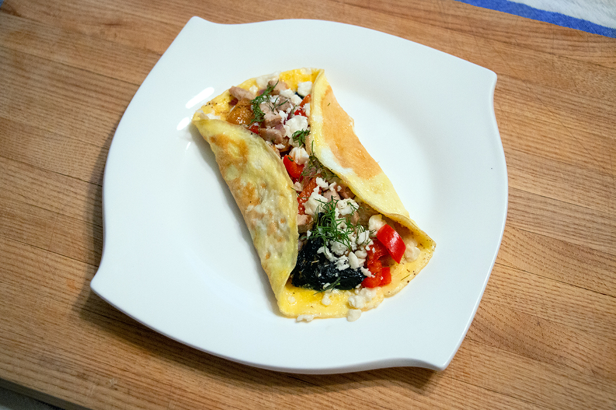White plate on a wooden cutting board, topped with an omelette filled with meat, peppers and feta cheese.