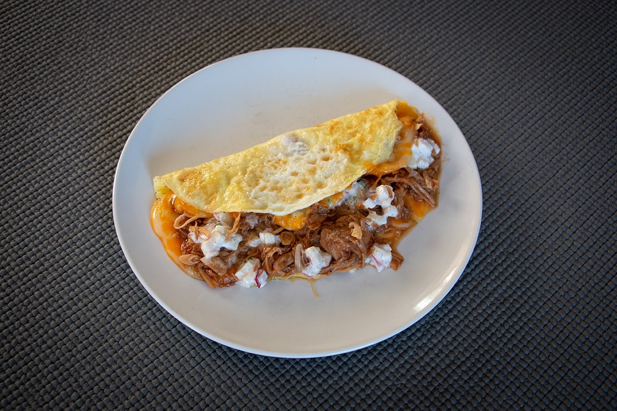 White plate with an omelette stuffed with cheese, pulled pork and a white sauce.