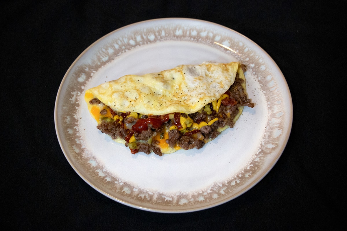 White and brown plate with an omelette stuffed with cheeseburger filling.