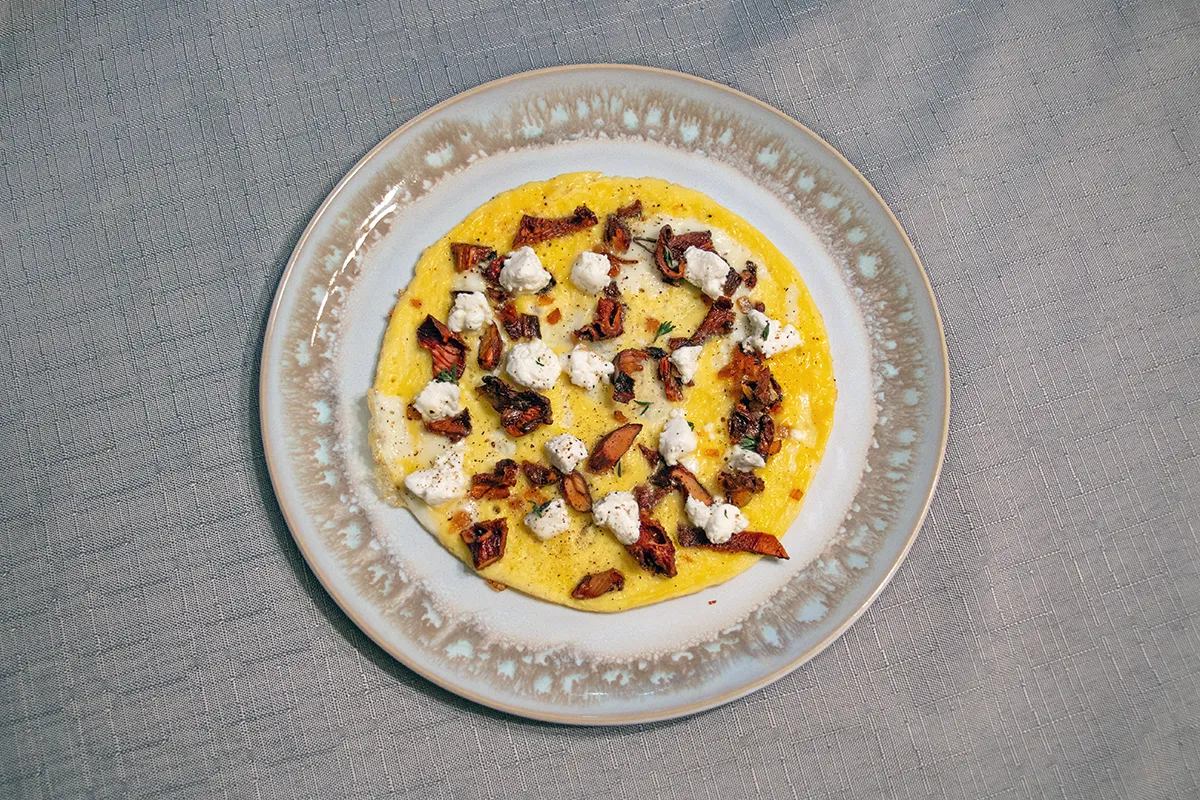 White and brown plate with an open-faced omelette on it. It is topped with chanterelle mushrooms, goat cheese and fresh thyme.