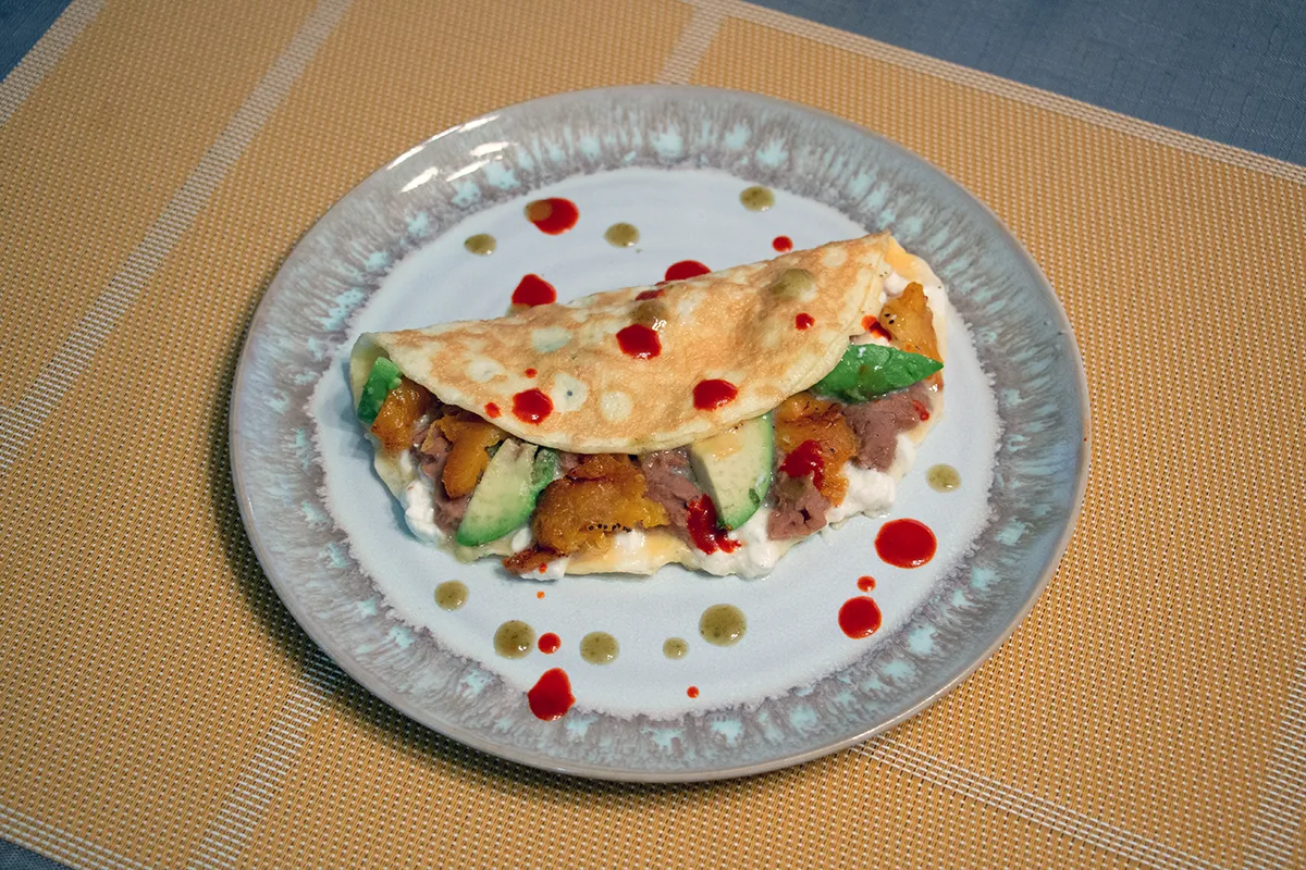 White and grey plate on a yellow placemat. On the plate is an omelette filled with beans, cheese, avocado and plantain.