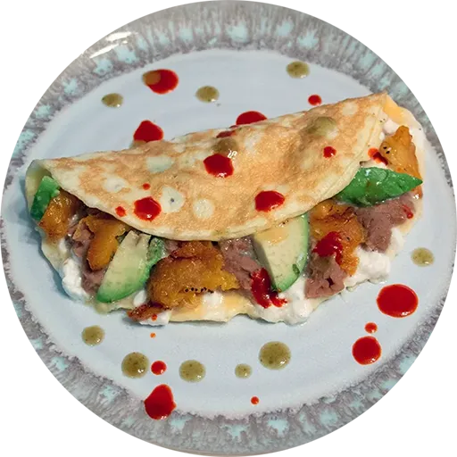 White and grey plate on a yellow placemat. On the plate is an omelette filled with beans, cheese, avocado and plantain.
