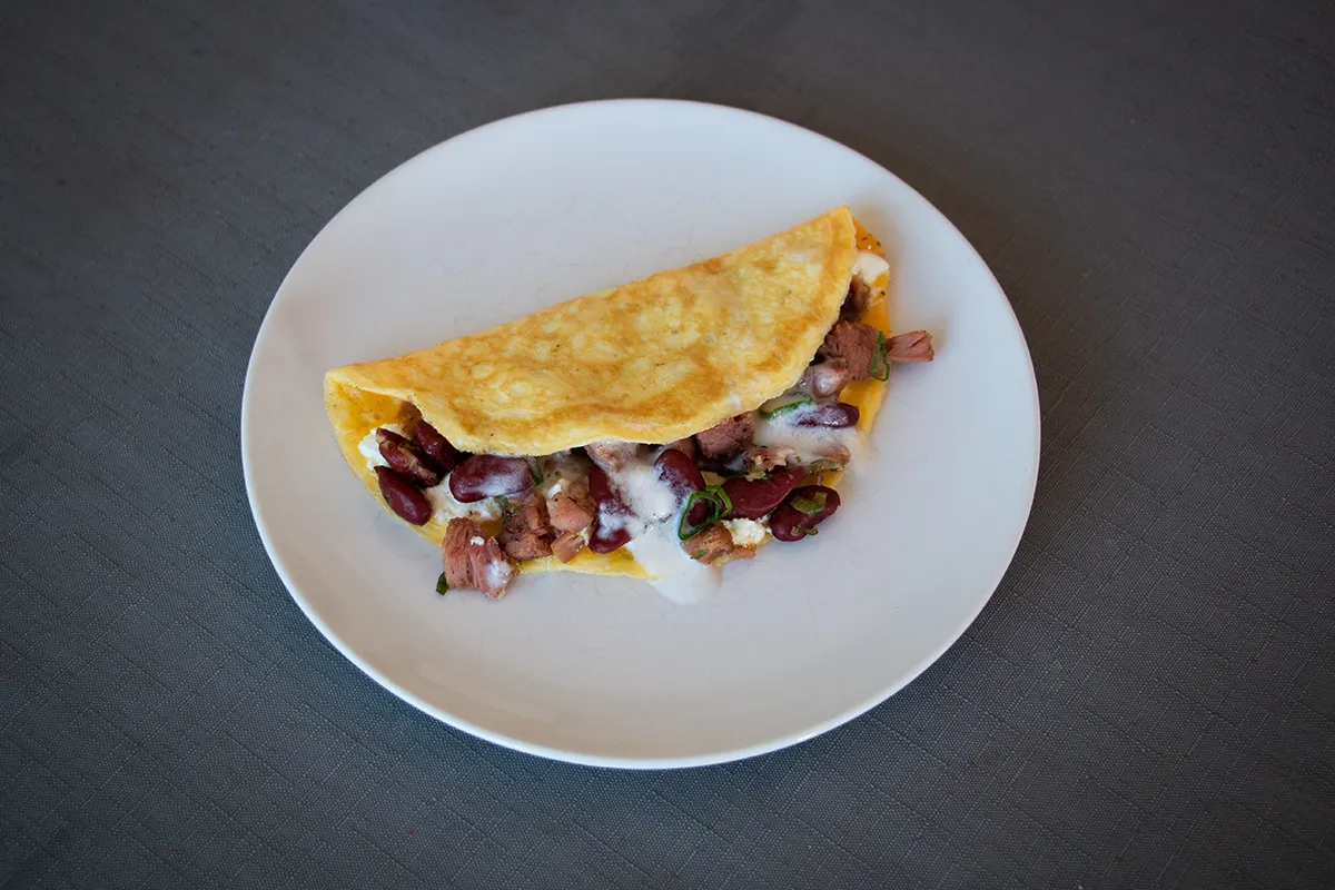 White plate with an omelette filled with ham, red beans, and a white coconut sauce.