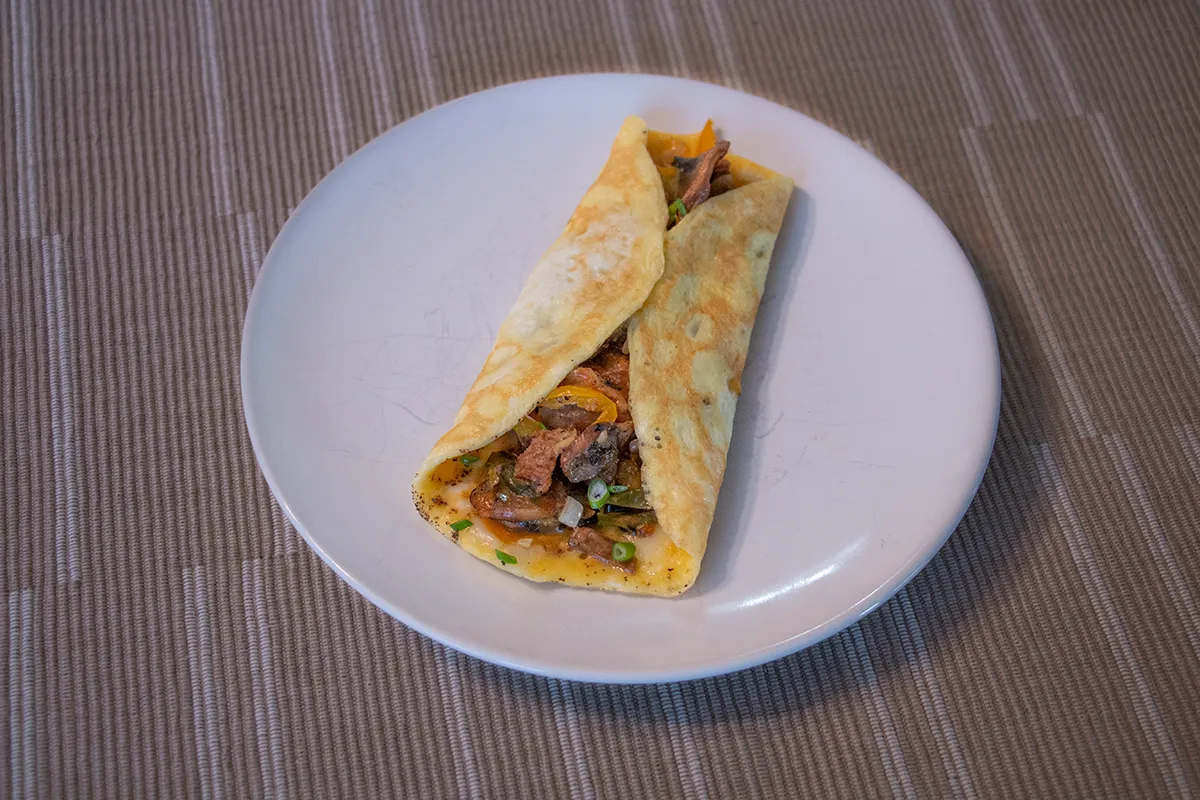 White plate with a rolled omelette filled with beef, onions, peppers, mushrooms and cheese.