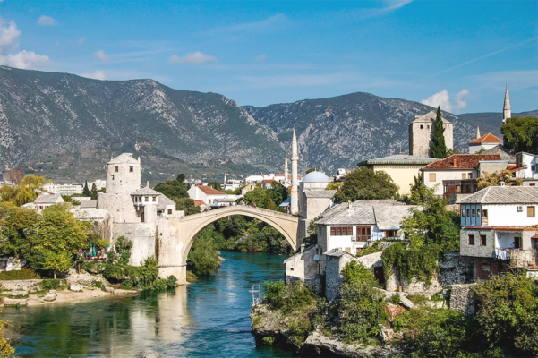 A village of wood and stone buildings, with several tall spires, along a river. A large arched bridge crosses the river.