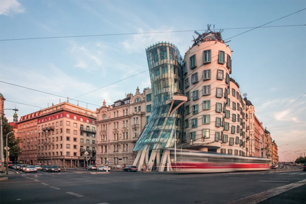 A build of strange architecture along a street. One side is twisted and surrounded by glass, the other is cylindrical. A red tram passes by.