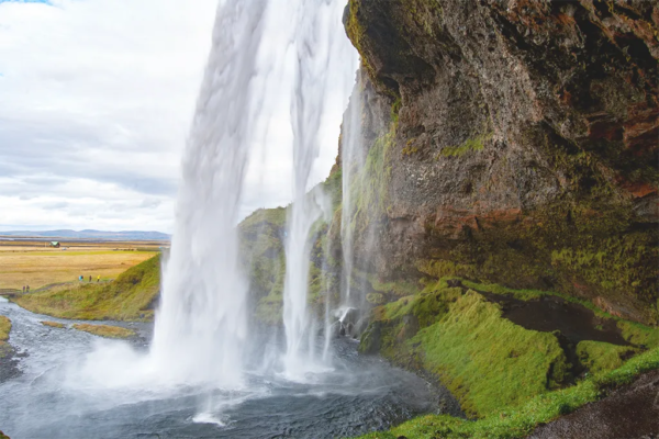 Looking through a waterfall from behind. Vibrant green grass grows below the rocky ledge behind the falls.