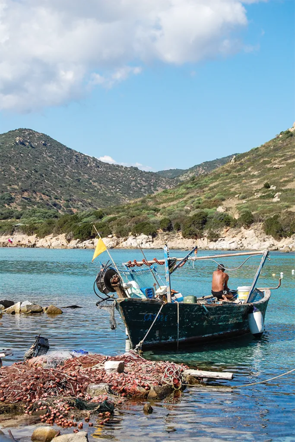 A man sits in a fishing boat in calm aqua-coloured water. Green hills line the shore.