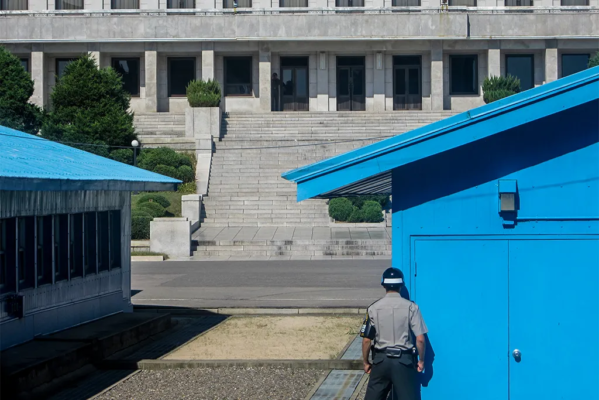 A guard stares towards a large stone building while standing beside a small blue building.