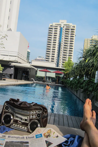 Rooftop swimming pool with a woman standing in it. In the foreground is a beach chair with a purse and a newspaper.